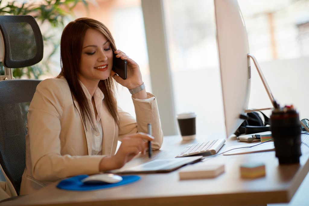Eine lächelnde Frau in Geschäftskleidung sitzt an ihrem Homeoffice-Schreibtisch, telefoniert und schreibt auf einem Notizblock. Vor ihr stehen ein Computer, eine Kaffeetasse und Büromaterialien - ein Beispiel dafür, wie man bei COVID-19 richtig arbeitet.
