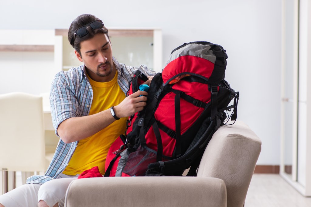 Ein Mann sitzt auf einer Couch, tr&auml;gt ein gelbes Hemd und eine Sonnenbrille auf dem Kopf und packt einen gro&szlig;en rot-schwarzen Rucksack in ein helles, modernes Wohnzimmer.