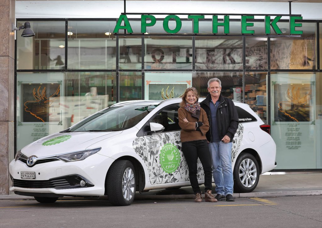 Eine lächelnde Frau und ein lächelnder Mann stehen neben einem weißen Auto mit grünen und floralen Motiven und werben für ihren Hauslieferservice, der vor einem Gebäude mit einem großen APOTHEKE-Schild über dem Eingang geparkt ist.