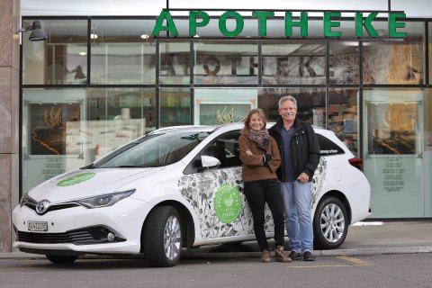 Eine l&auml;chelnde Frau und ein l&auml;chelnder Mann stehen neben einem wei&szlig;en Auto mit gr&uuml;nen und floralen Motiven und werben f&uuml;r ihren Hauslieferservice, der vor einem Geb&auml;ude mit einem gro&szlig;en APOTHEKE-Schild &uuml;ber dem Eingang geparkt ist.