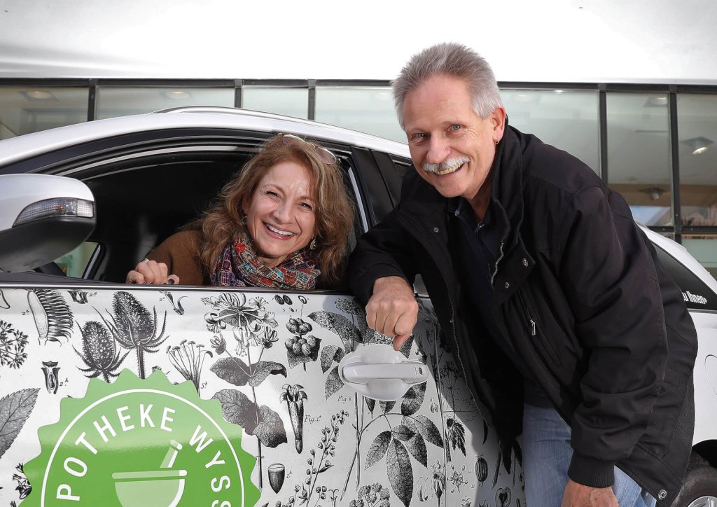 Eine l&auml;chelnde Frau sitzt in einem mit botanischen Illustrationen geschm&uuml;ckten Auto, das einen freundlichen Heimservice darstellt, w&auml;hrend ein l&auml;chelnder Mann neben ihr steht und sich an das offene Fenster lehnt. Beide sehen gl&uuml;cklich und einladend aus.