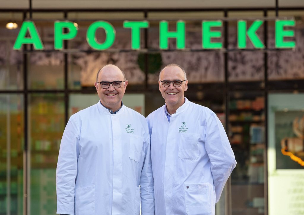 Zwei lächelnde Männer in weißen Laborkitteln stehen vor einem Gebäude mit einem großen grünen Apothekenschild, das auf ihre Stammapotheke hinweist. Sie scheinen Apotheker oder Angestellte zu sein.