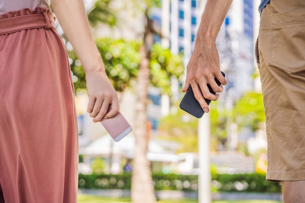 Zwei Personen stehen im Freien nebeneinander und halten jeweils ein Smartphone in der Hand - perfekt, um die SwissCovid App oder andere Tools zur Kontaktsuche zu nutzen. Die linke Person trägt einen pinken Rock und hält ein pinkes Telefon, die rechte eine hellbraune Shorts und ein schwarzes Telefon.