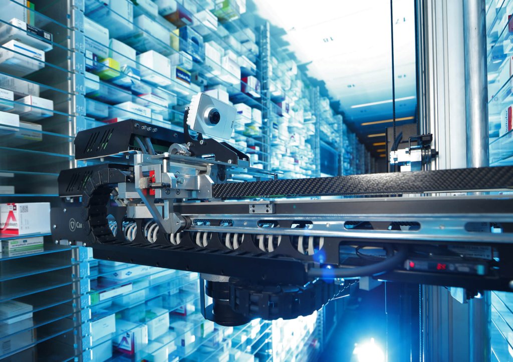 Ein Roboterarm entnimmt Medikamentenboxen in einem automatischen Apothekenlagersystem und sorgt für eine korrekte Lagerung. Sauber geordnete Reihen von Arzneimitteln werden durch helle, blau getönte Beleuchtung für eine optimale Medikamentenaufbewahrung beleuchtet.