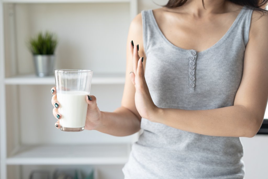 Eine Frau in einem grauen Tank-Top h&auml;lt ein Glas Milch in der einen Hand und macht mit der anderen Hand eine abweisende Geste, die Symptome einer Laktoseunvertr&auml;glichkeit andeutet.