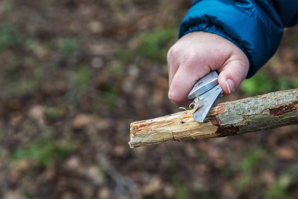 Eine Person in einer blauen Jacke schnitzt mit einem Klappmesser einen Stock im Freien, w&auml;hrend ihre mobile Mietapotheke in der N&auml;he auf dem unscharfen Waldboden im Hintergrund ruht.