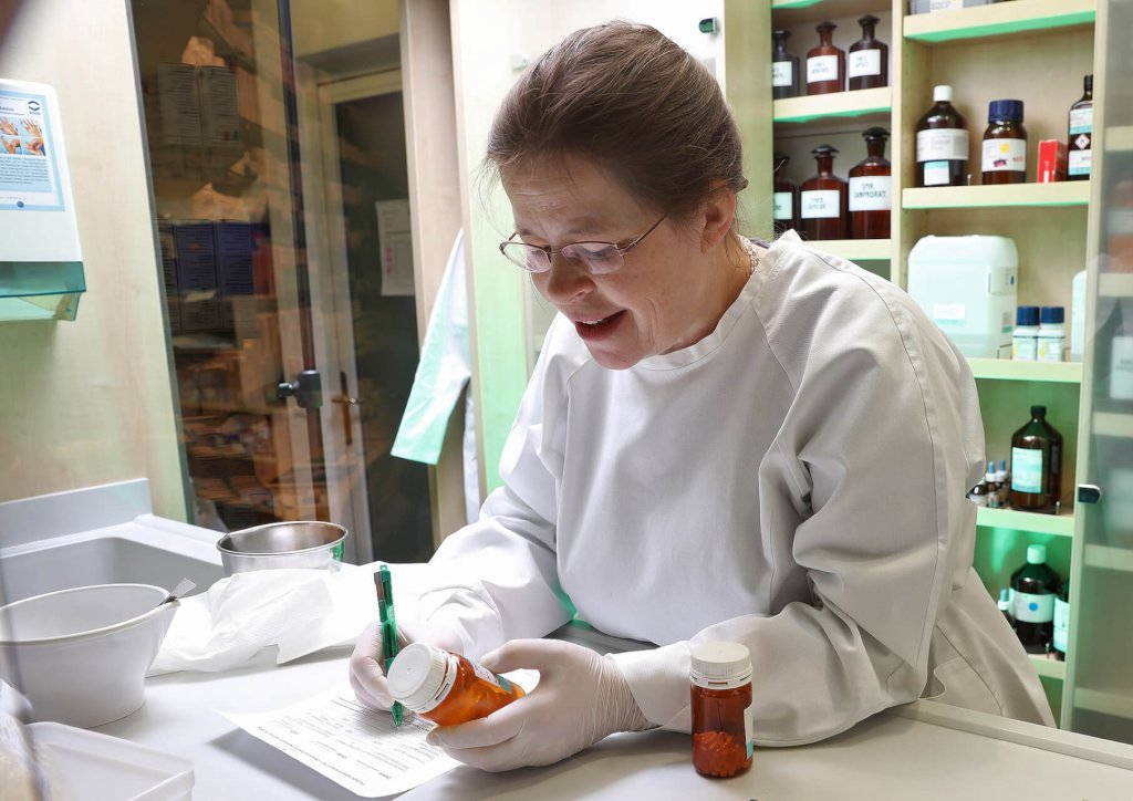 Eine Apothekerin im weißen Kittel und mit Handschuhen etikettiert eine Rezeptflasche an einem Apothekentresen, umgeben von Arzneimittelflaschen, Papierkram und QMS-Dokumenten. Sie lächelt und konzentriert sich auf ihre Aufgabe.