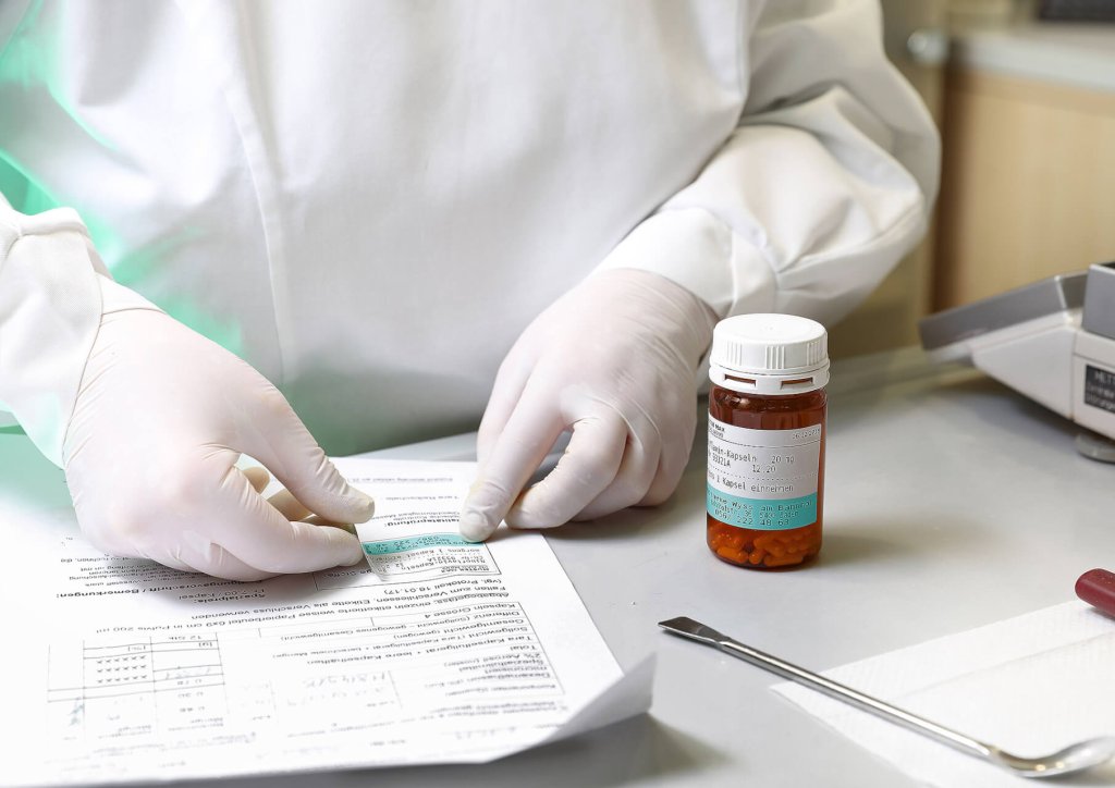 Eine Person mit wei&szlig;en Handschuhen pr&uuml;ft ein medizinisches Dokument im Rahmen eines Qualit&auml;tsmanagementprozesses (QMS) und versieht es mit einem Etikett. Eine braune, verschreibungspflichtige Pillenflasche und ein Metallinstrument liegen auf dem Tisch daneben.
