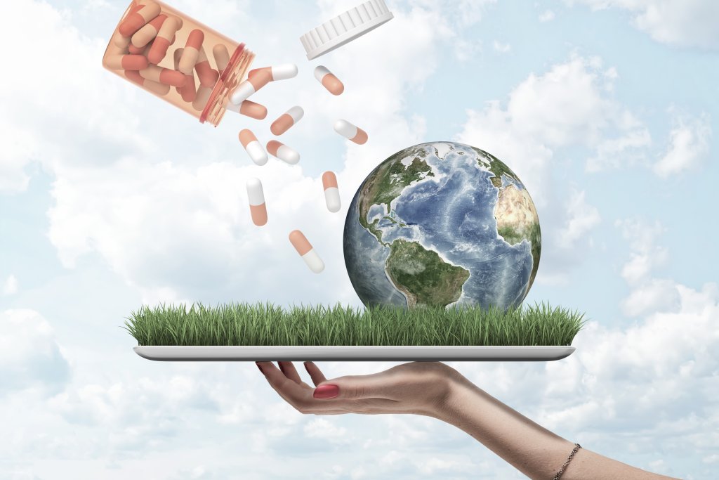 Eine Hand h&auml;lt ein Tablett mit Gras, einen Globus und Pillen, die aus einer offenen Pillenflasche fallen, vor einem wolkenverhangenen Himmel - als Symbol daf&uuml;r, wie Antibiotika und ihre Verwendung die Welt und die Umwelt beeinflussen und zur Antibiotikaresistenz beitragen.