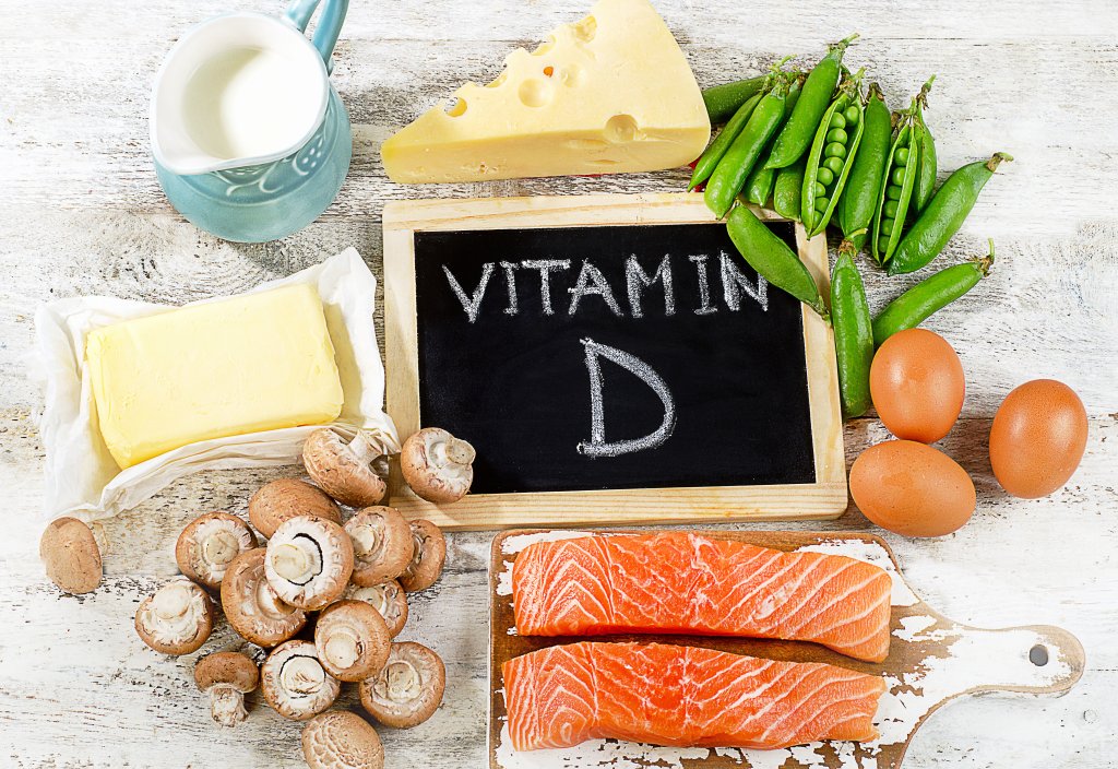 Eine Vielzahl von Vitamin-D-reichen Lebensmitteln, darunter Lachs, Eier, Pilze, K&auml;se, Erbsen, Butter und ein Krug Milch, sind um eine kleine Kreidetafel herum angeordnet, auf der VITAMIN D-perfekt f&uuml;r gesund durch den Winter und nat&uuml;rlich stark bleiben steht.
