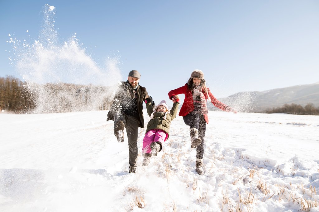 Eine dreiköpfige Familie genießt einen natürlichen Wintertag und spielt im Schnee. Die Eltern schwingen ihr Kind zwischen sich, alle lächeln in warmer Kleidung, während der Schnee herumfliegt. Hügel und Bäume vervollständigen die gesunde Outdoor-Szene.