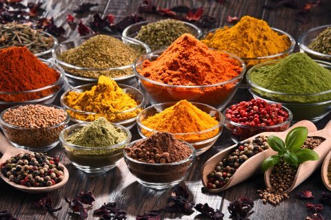 Eine Vielzahl bunter Gew&uuml;rze und Kr&auml;uter, die als Heilmittel gesch&auml;tzt werden, sind in kleinen Glassch&auml;lchen und Holzl&ouml;ffeln auf einer rustikalen Holzfl&auml;che ausgestellt, darunter Pulver und ganze Samen in Rot-, Gelb-, Gr&uuml;n- und Braunt&ouml;nen.