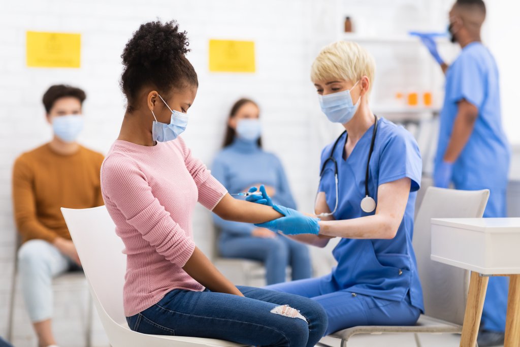 Eine Krankenschwester in blauem Kittel bereitet den Arm einer jungen Frau für eine Grippeimpfung vor. Beide tragen Masken. Im Hintergrund warten drei weitere maskierte Personen in einem hell erleuchteten Raum auf ihren Einsatz.