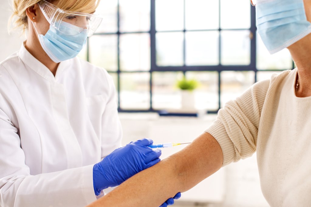 Ein Mitarbeiter des Gesundheitswesens, der eine Maske, eine Schutzbrille und Handschuhe tr&auml;gt, verabreicht einem Patienten, der ebenfalls eine Maske tr&auml;gt, in einer hellen medizinischen Umgebung eine Impfung gegen Grippeimpfung in den Oberarm.