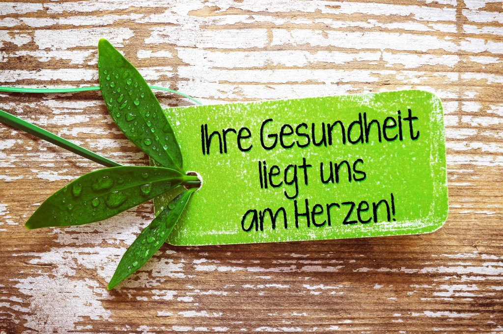 Ein grünes Schild mit Wassertropfen und Blättern mit der Aufschrift Ihre Gesundheit liegt uns am Herzen! auf einer rustikalen Holzoberfläche unterstreicht die Bedeutung von Gesundheit und Wohlbefinden - Ihre Gesundheit liegt uns am Herzen!.