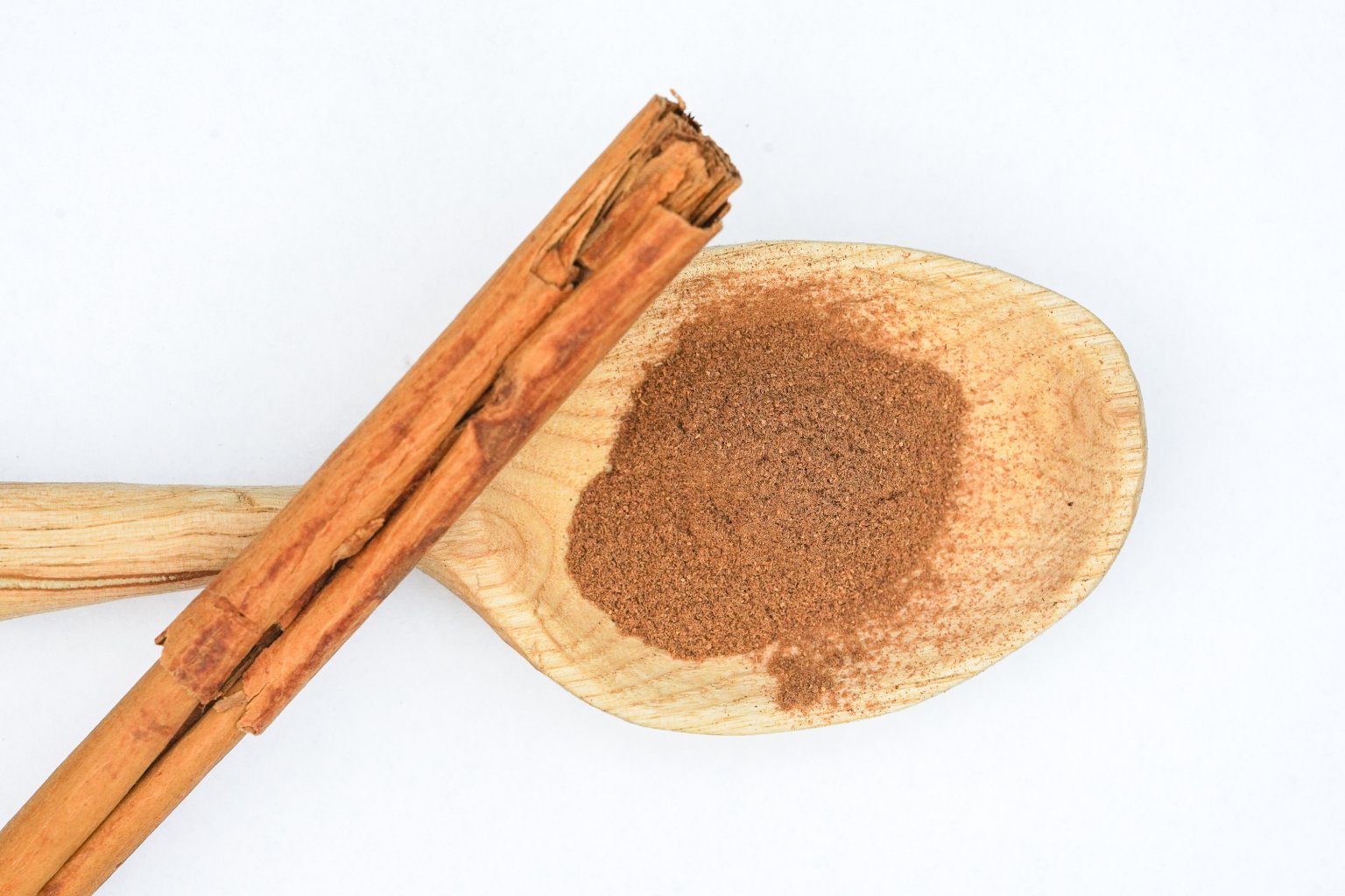 Ein Holzl&ouml;ffel h&auml;lt einen Haufen gemahlenen Zimt, ein Klassiker unter den Gew&uuml;rzen und Heilmitteln, w&auml;hrend eine ganze Zimtstange daneben liegt, beide auf wei&szlig;em Hintergrund.