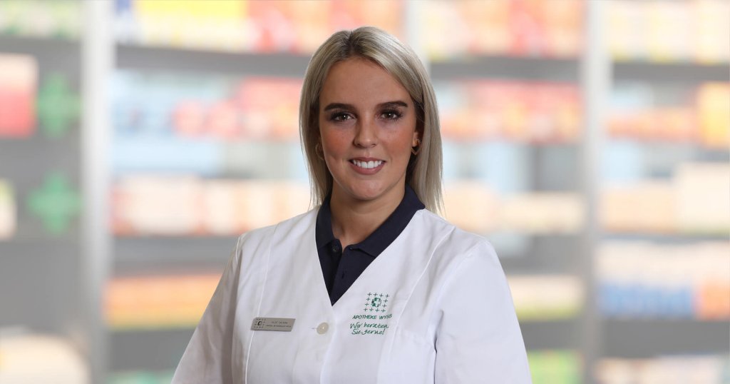 Alice Troisi, die einen weißen Laborkittel und ein Namensschild trägt, steht lächelnd vor unscharfen Regalen, die auf eine Apotheke oder einen Gesundheitsbereich hindeuten.