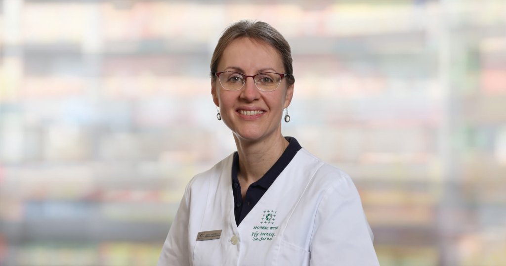 Anita Zimmermann, die eine Brille und einen weißen Laborkittel mit einem Namensschild trägt, lächelt in die Kamera. Der Hintergrund ist unscharf und farbenfroh, was auf eine Innenraumumgebung hindeutet.