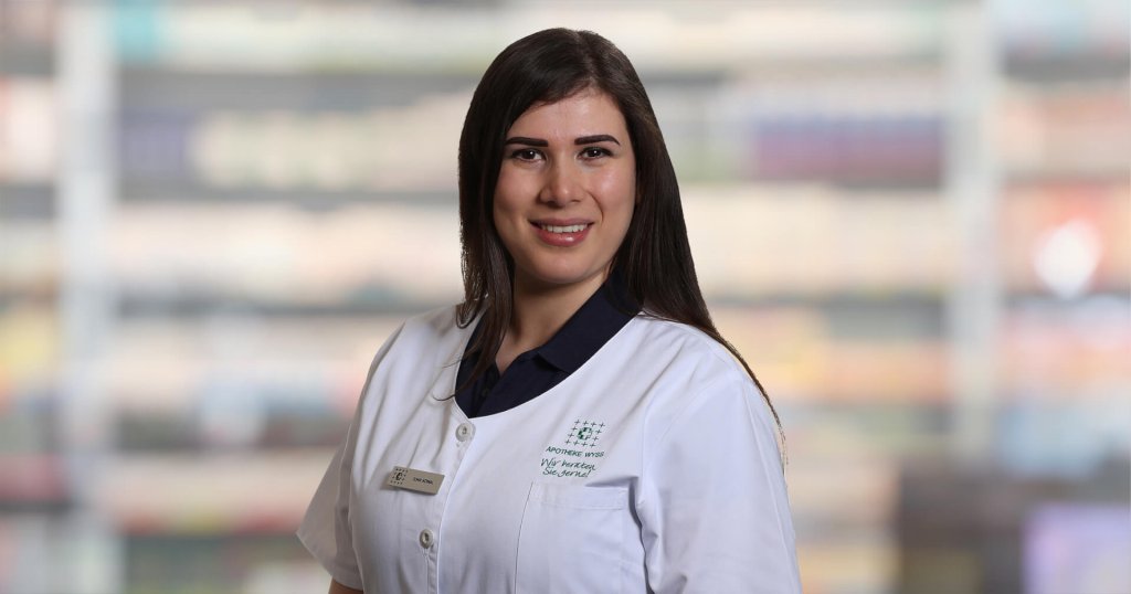 Bermal Demir, eine Frau mit langen dunklen Haaren, die einen weißen Laborkittel und ein Namensschild trägt, steht lächelnd vor verschwommenen Regalen, wahrscheinlich in einer Apotheke oder einem medizinischen Bereich.