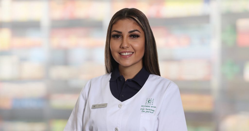Joana Lourenco, die einen weißen Apothekenkittel und ein dunkles Hemd trägt, steht lächelnd vor verschwommenen Regalen. Sie hat langes braunes Haar, ein Namensschild und scheint sich in einer Apotheke oder einem Geschäft zu befinden.