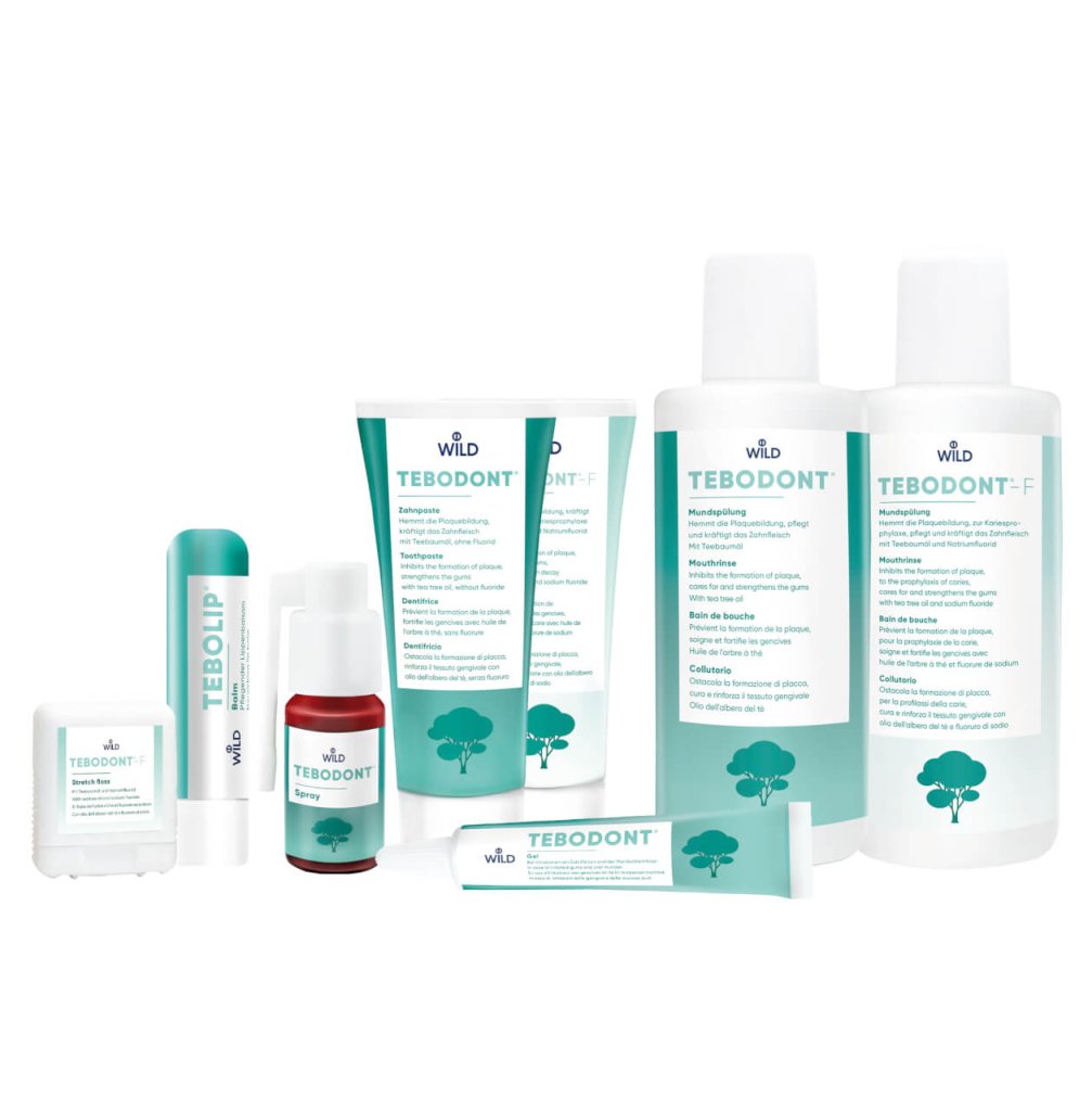 Eine Kollektion von Wild TEBODONT-Zahnpflegeprodukten, einschlie&szlig;lich Zahnpasta, Mundwasser, Zahnseide, Lippenbalsam und Mundsp&uuml;lung, mit wei&szlig;en und blaugr&uuml;nen Verpackungen mit Baumsymbolen.