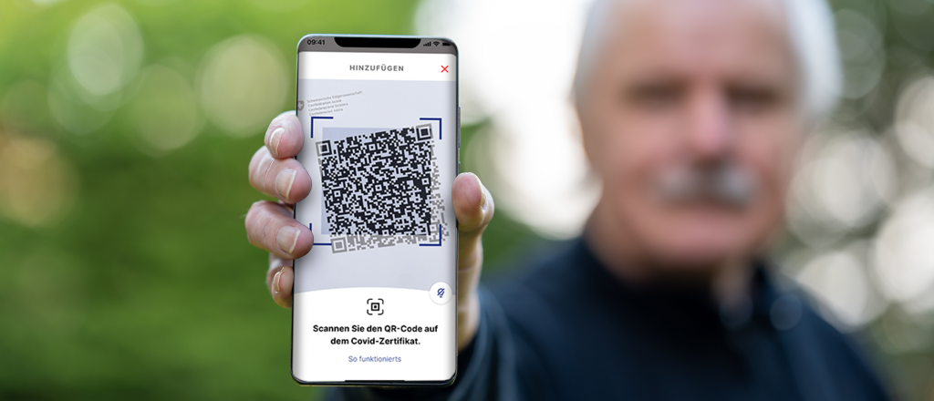 Eine Person hält ein Smartphone in der Hand, auf dem ein großer QR-Code angezeigt wird, wahrscheinlich für ein COVID-Zertifikat, mit deutschem Text unter dem Code. Der Hintergrund ist unscharf und zeigt einen älteren Mann.