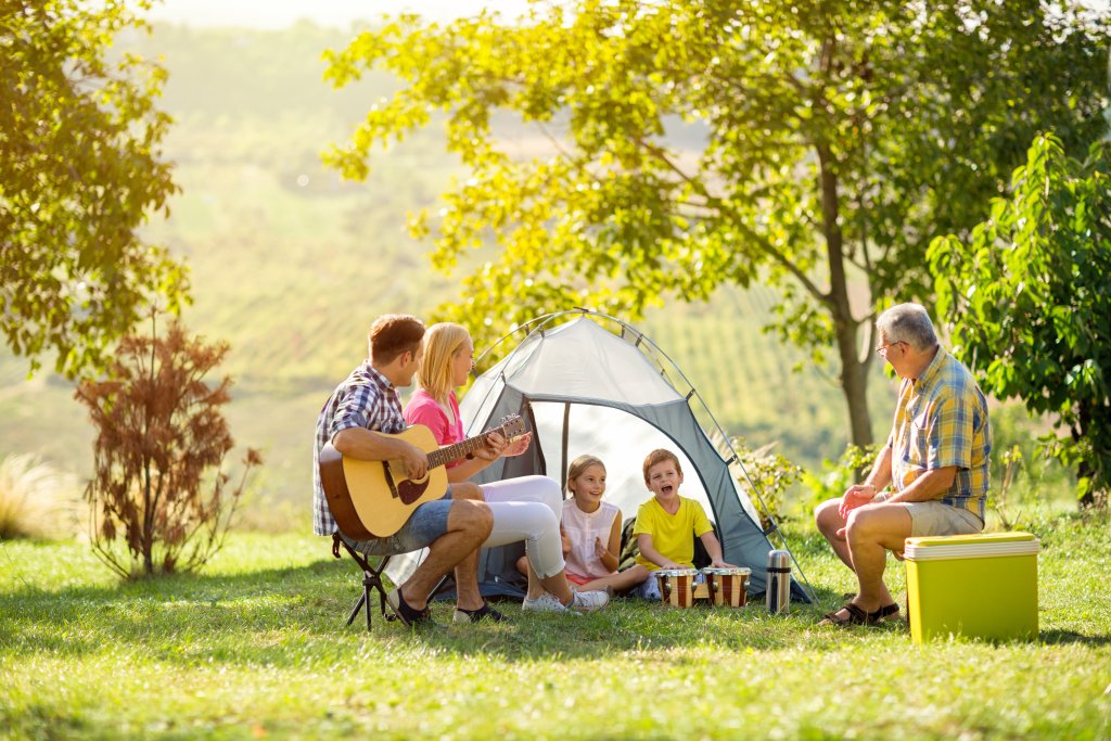 Eine Familie und ein älterer Mann sitzen mit ihrem Outdoorspass an einem Zelt im Freien. Der Mann spielt Gitarre, zwei Kinder spielen Schlagzeug, und alle lächeln und genießen den sonnigen Tag inmitten von grünen Bäumen und Gras.