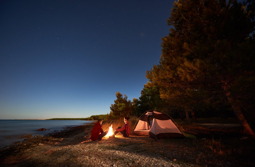 Zwei Menschen sitzen nachts an einem Lagerfeuer in der N&auml;he eines Zeltes an einem felsigen Seeufer, umgeben von B&auml;umen und unter einem Sternenhimmel. Mit einem Outdoorspass k&ouml;nnen Sie dieses heitere Wildnisabenteuer selbst erleben.