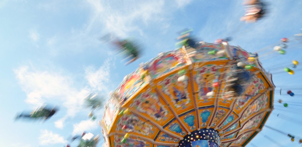 Eine farbenfrohe, sich drehende Karussellfahrt in einem Vergnügungspark, aufgenommen mit Bewegungsunschärfe vor einem blauen Himmel mit leichten Wolken, ruft Aufregung und den schwindelerregenden Rausch des Schwindels hervor.