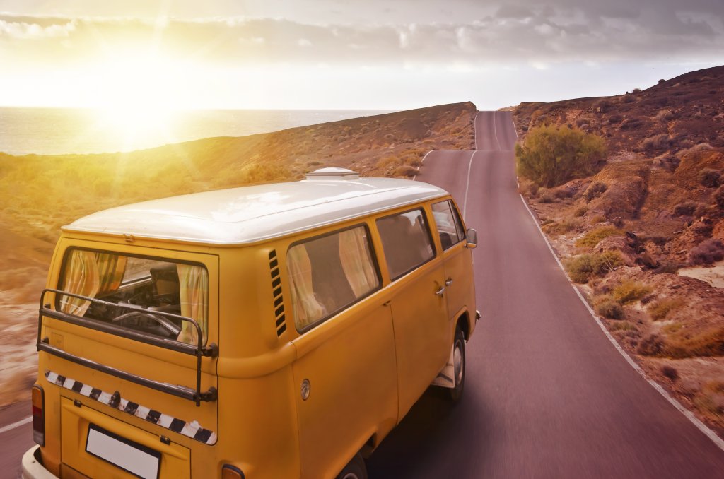 Ein gelber Oldtimer-Van f&auml;hrt auf einer leeren, gewundenen Stra&szlig;e durch eine W&uuml;stenlandschaft bei Sonnenuntergang, w&auml;hrend das Sonnenlicht den Himmel erhellt - perfekt f&uuml;r ein Outdoorspass-Abenteuer. Warme Farbt&ouml;ne verleihen dieser ruhigen Szene einen nostalgischen Glanz.