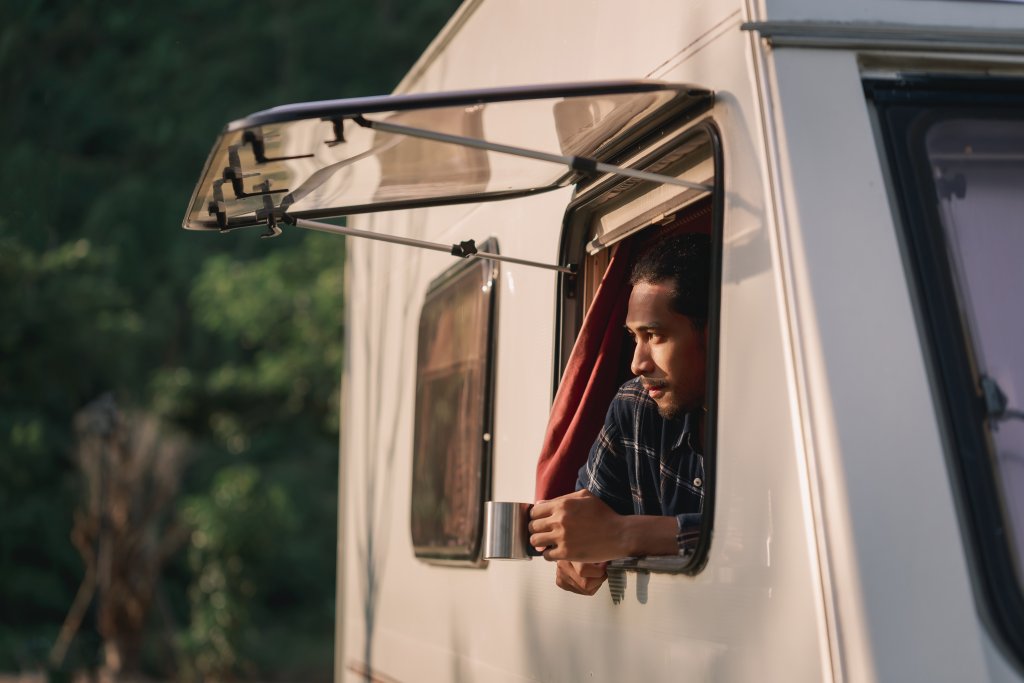 Ein Mann lehnt sich aus dem Fenster eines Wohnmobils, h&auml;lt einen Becher in der Hand und schaut nachdenklich in die Ferne. Mit seinem Outdoorspass im Schlepptau genie&szlig;t er die Ruhe der Natur, umgeben von &uuml;ppig gr&uuml;nem Laub.