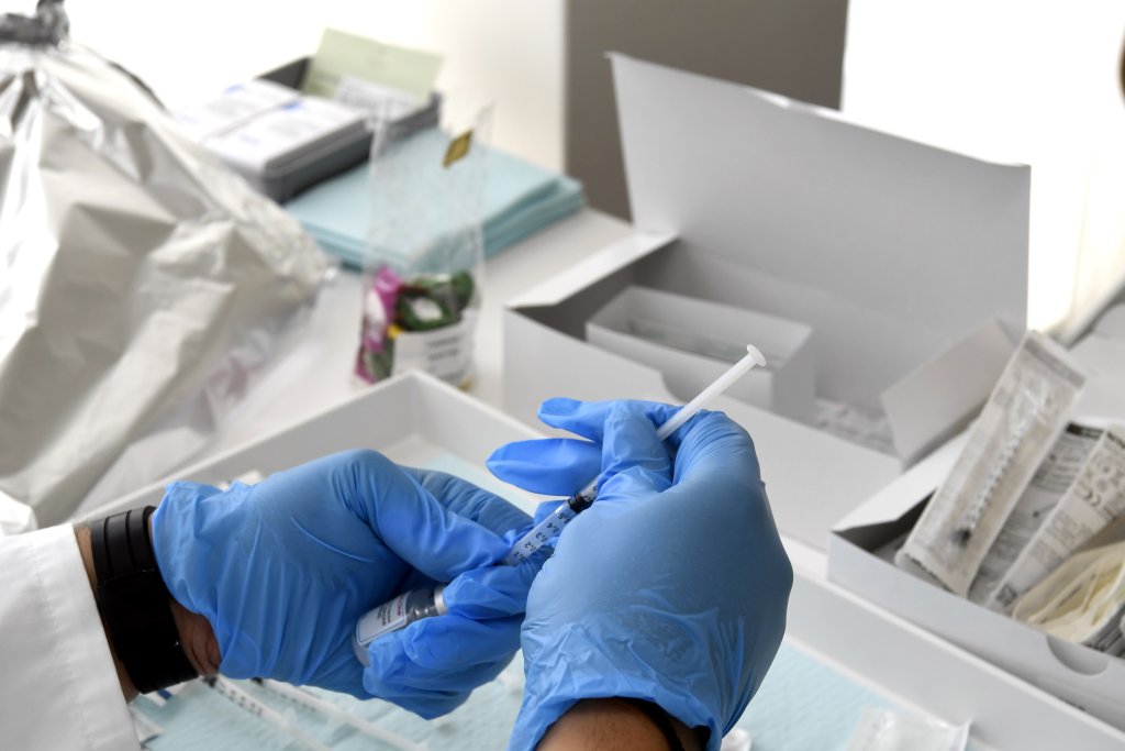 Eine Person mit blauen Handschuhen bereitet w&auml;hrend eines DGS-Besuchs eine Spritze vor, umgeben von medizinischem Material und Verpackungen auf einem Tisch in einer klinischen Umgebung.