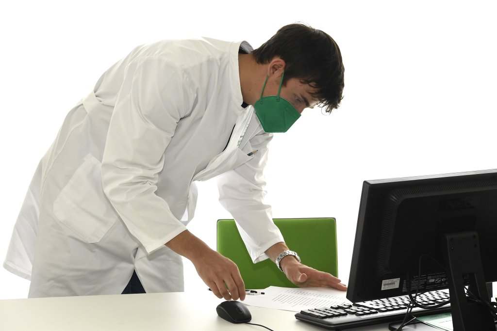 Eine Person in wei&szlig;em Laborkittel und gr&uuml;ner Gesichtsmaske steht w&auml;hrend eines DGS-Besuchs an einem Schreibtisch und pr&uuml;ft ein Dokument neben einem Computermonitor und einer Maus, mit einem gr&uuml;nen Stuhl im Hintergrund.