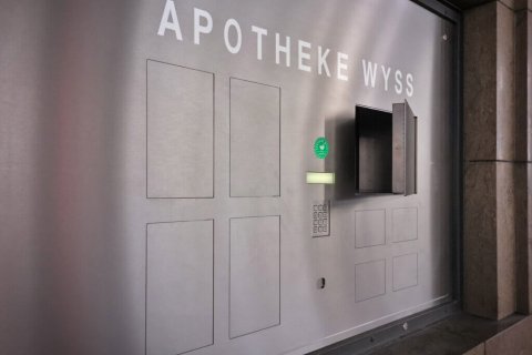 Ein metallisches Wandpaneel mit der Aufschrift APOTHEKE WYSS besteht aus mehreren rechteckigen Abschnitten, einer Tastatur, einem Display, einem gr&uuml;nen Aufkleber und einem offenen Fach - wahrscheinlich 24-Stunden-Abholf&auml;cher als Teil eines automatischen Apothekensystems.