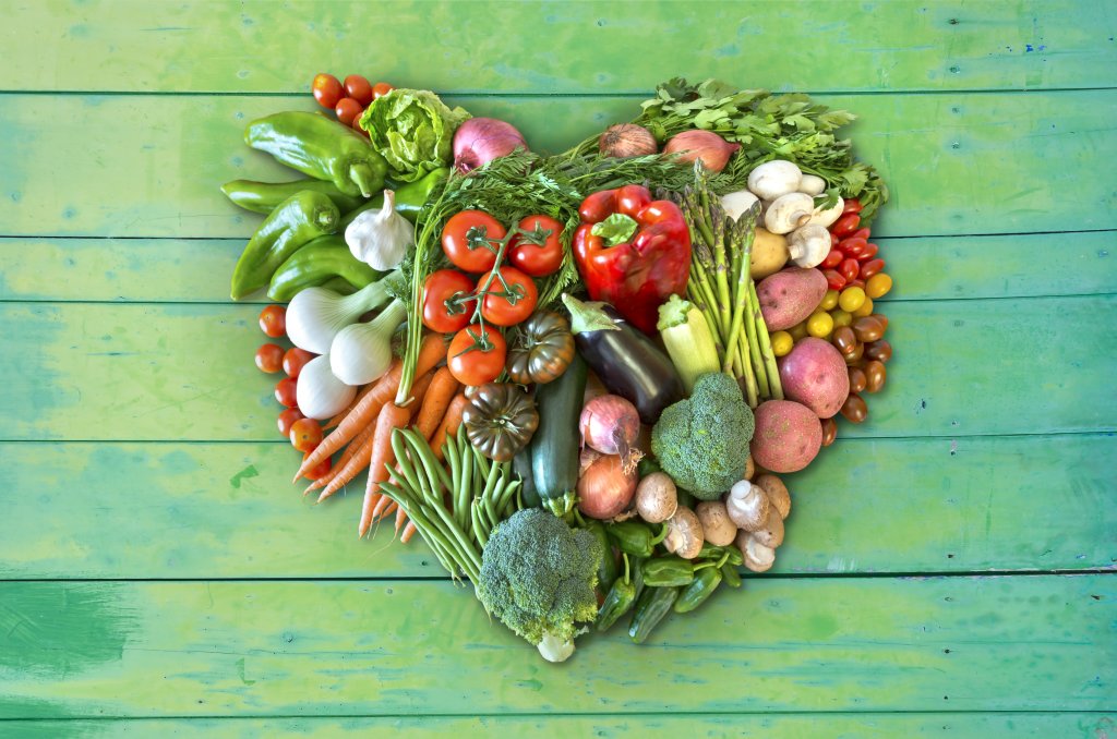 Eine Vielzahl von frischem Gem&uuml;se, darunter Tomaten, Paprika, Brokkoli, Zwiebeln und Pilze - lebenswichtig f&uuml;r Ihr Wohlbefinden - sind in Form eines Herzens auf einer gr&uuml;nen Holzfl&auml;che angeordnet.