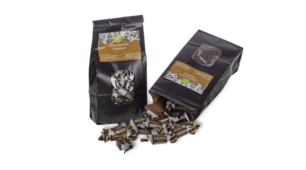 Zwei schwarze T&uuml;ten mit Lakritzbonbons, von denen eine aufrecht steht und die andere auf der Seite liegt, so dass die Bonbons herausquellen. Die Bonbons sind einzeln in schwarzes und braunes Papier eingewickelt.