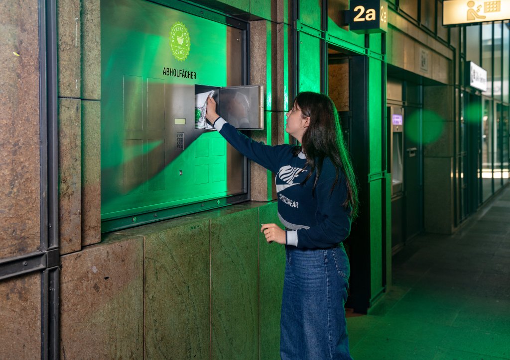 Eine Frau steht vor einem Geb&auml;ude und holt ein Paket aus einem Schlie&szlig;fachfenster von 24-Std-Abholf&auml;cher mit der Aufschrift ABHOLFL&Auml;CHE in einem gr&uuml;n beleuchteten Bereich. Sie tr&auml;gt einen blauen Pullover und Jeans.