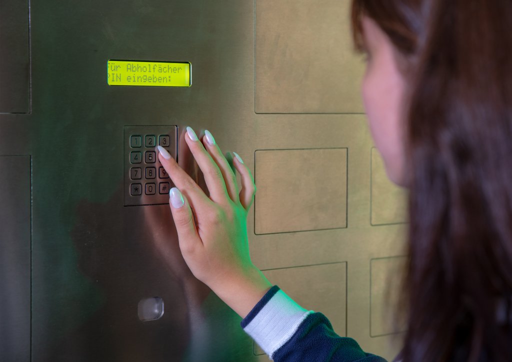 Eine Person gibt einen Code auf einem Tastenfeld neben einer digitalen Anzeige mit deutschem Text ein, die an einem metallischen 24-Std-Abholf&auml;cher-System angebracht ist. Auf dem Display steht Im Abholf&auml;chern IN eingegeben.
