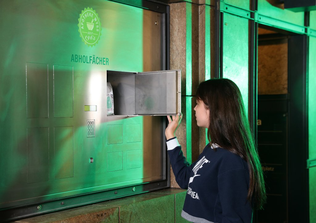 Ein junges M&auml;dchen &ouml;ffnet ein 24-Std-Abholf&auml;cher-Metallfach mit der Aufschrift Abholf&auml;cher, um ein Paket herauszunehmen. Das in eine Wand eingelassene und gr&uuml;n beleuchtete Schlie&szlig;fach ist dem M&auml;dchen zugewandt, das in einem dunklen Pullover steht.