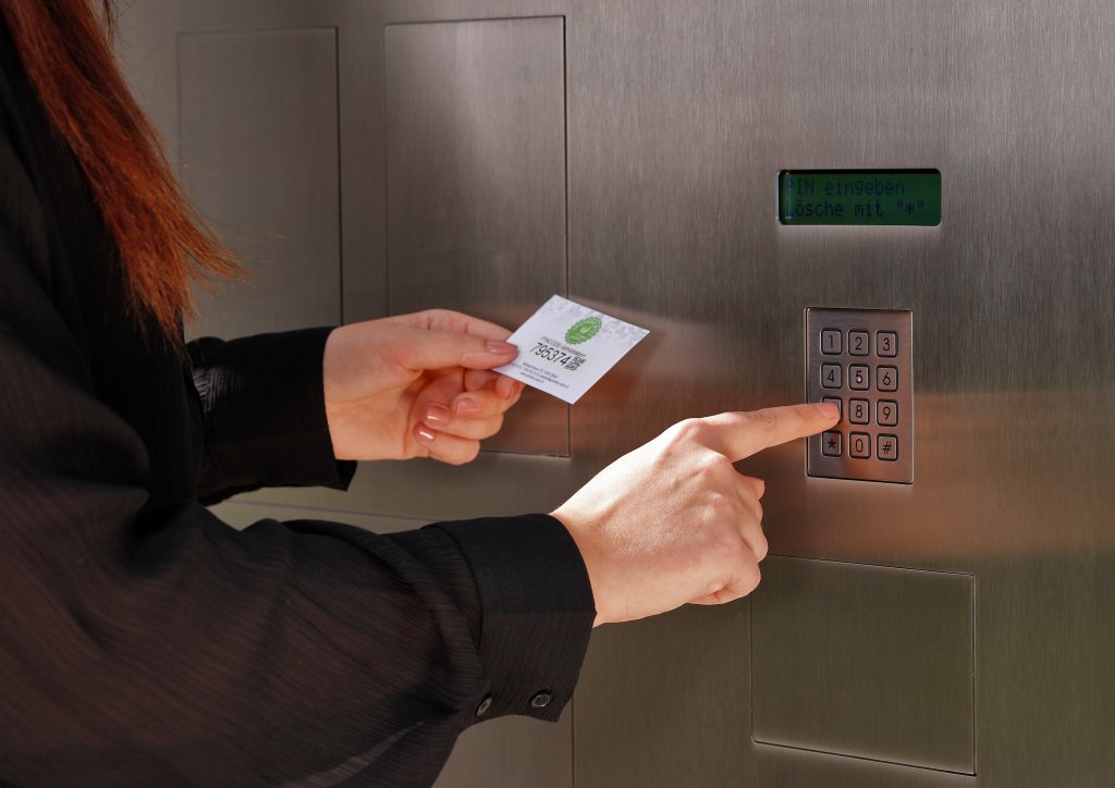 Eine Person, die eine Karte in der Hand h&auml;lt, gibt einen Code auf einem Tastenfeld ein, das an einer Edelstahlwand angebracht ist, und erh&auml;lt so Zugang zu sicheren 24-Std-Abholf&auml;chern.