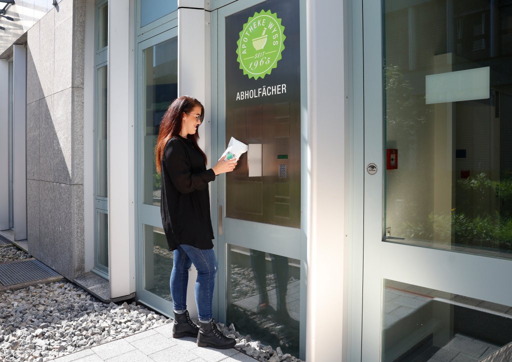 Eine Frau steht vor einem Geb&auml;ude und h&auml;lt ein Paket vor Schlie&szlig;f&auml;chern mit einem gr&uuml;nen Schild, auf dem Apotheke seit 1683 24-Std-Abholf&auml;cher steht. Sie scheint das Paket zu holen oder abzulegen.