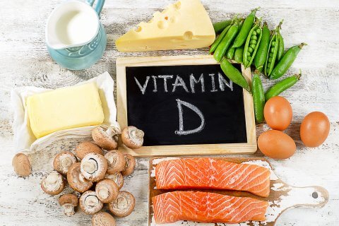 Eine Kreidetafel mit der Aufschrift Vitamin D, auch bekannt als Sonnenvitamin, ist umgeben von Lachsfilets, Eiern, Butter, K&auml;se, Pilzen, gr&uuml;nen Erbsen und einem Krug Milch auf einer wei&szlig;en Holzfl&auml;che.