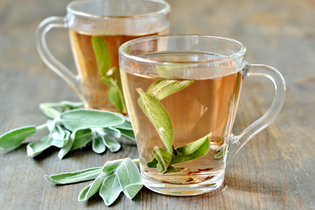 Zwei klare Glastassen mit Kr&auml;utertee und frischen Salbeibl&auml;ttern, einem bekannten Heilmittel, stehen auf einer Holzplatte. Weitere Salbeibl&auml;tter und Gew&uuml;rze sind neben den Tassen angeordnet. Der Tee leuchtet in einer hellen goldbraunen Farbe.