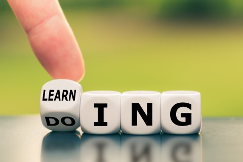 Eine Hand dreht einen W&uuml;rfel mit der Aufschrift DO um, so dass das Wort LEARN erscheint. So entsteht der Satz LEARNING from DOING, der das Wachstum in einem &Uuml;berbetrieblichen Kurs (&Uuml;K) symbolisiert. Die Bl&ouml;cke liegen auf einer reflektierenden Oberfl&auml;che mit einem verschwommenen gr&uuml;nen Hintergrund.