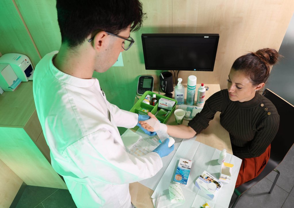 Eine medizinische Fachkraft in einem wei&szlig;en Kittel und mit blauen Handschuhen bereitet sich darauf vor, einer sitzenden Frau w&auml;hrend eines &Uuml;berbetrieblichen Kurses (&Uuml;K) in einer Klinik einen Verband anzulegen, w&auml;hrend zwischen ihnen auf einem Tisch medizinisches Material und Ger&auml;te liegen.