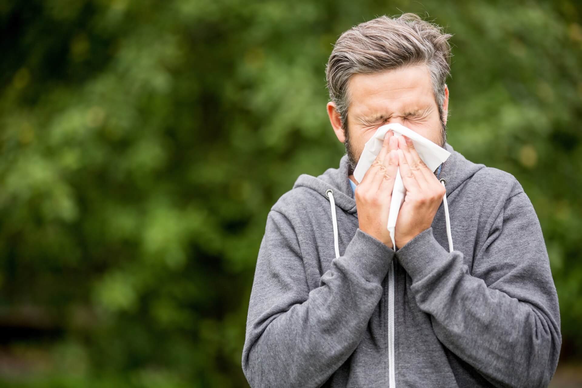 Ein Mann in einem grauen Kapuzenpulli steht im Freien und niest mit geschlossenen Augen in ein Taschentuch, wahrscheinlich hat er Heuschnupfen. Im Hintergrund ist unscharfes gr&uuml;nes Laub zu sehen.