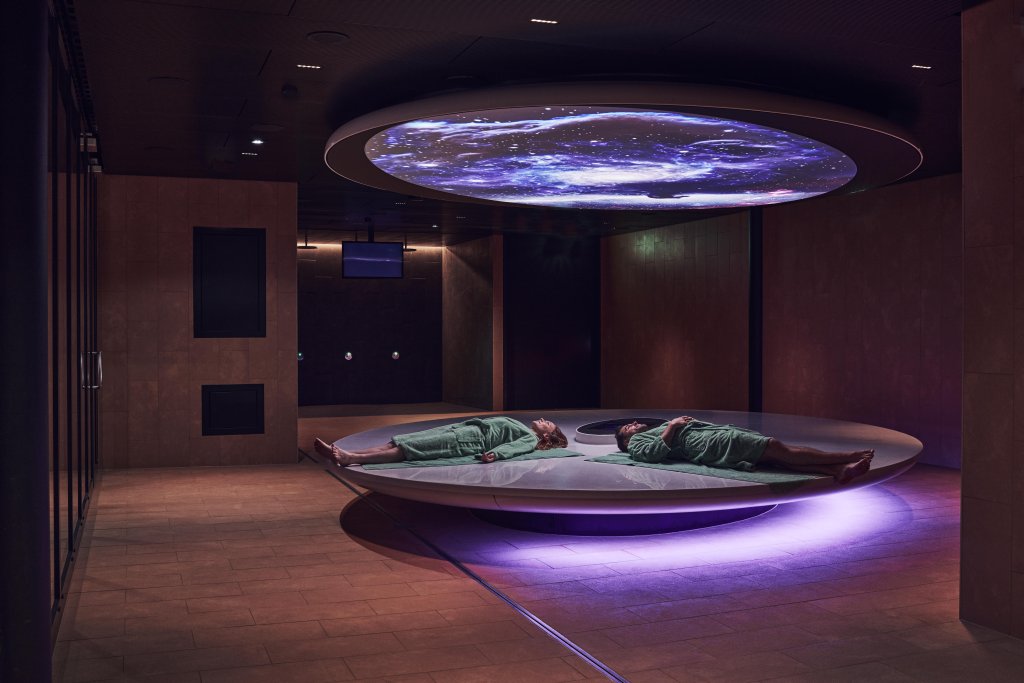 Zwei Personen in gr&uuml;nen Gew&auml;ndern liegen auf einem gro&szlig;en, runden Podest unter einem kosmischen Himmelsbild in einem schwach beleuchteten, modernen FORTYSEVEN-Entspannungsraum mit sanftem lila Licht - Wellness f&uuml;r die Sinne pur.