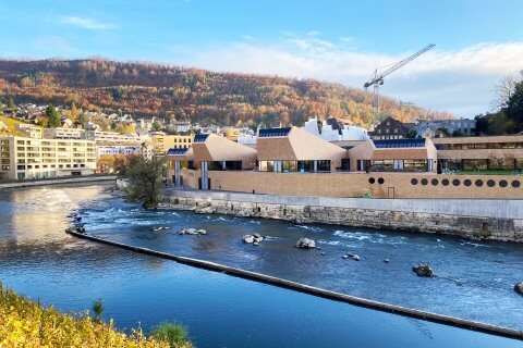 Moderne Backsteingeb&auml;ude mit kantigen D&auml;chern liegen an einem Fluss mit sanfter Str&ouml;mung; im Hintergrund bedecken Herbstb&auml;ume den Hang, und &uuml;ber der FORTYSEVEN-Wellness-Oase ist unter blauem Himmel ein Kran zu sehen.