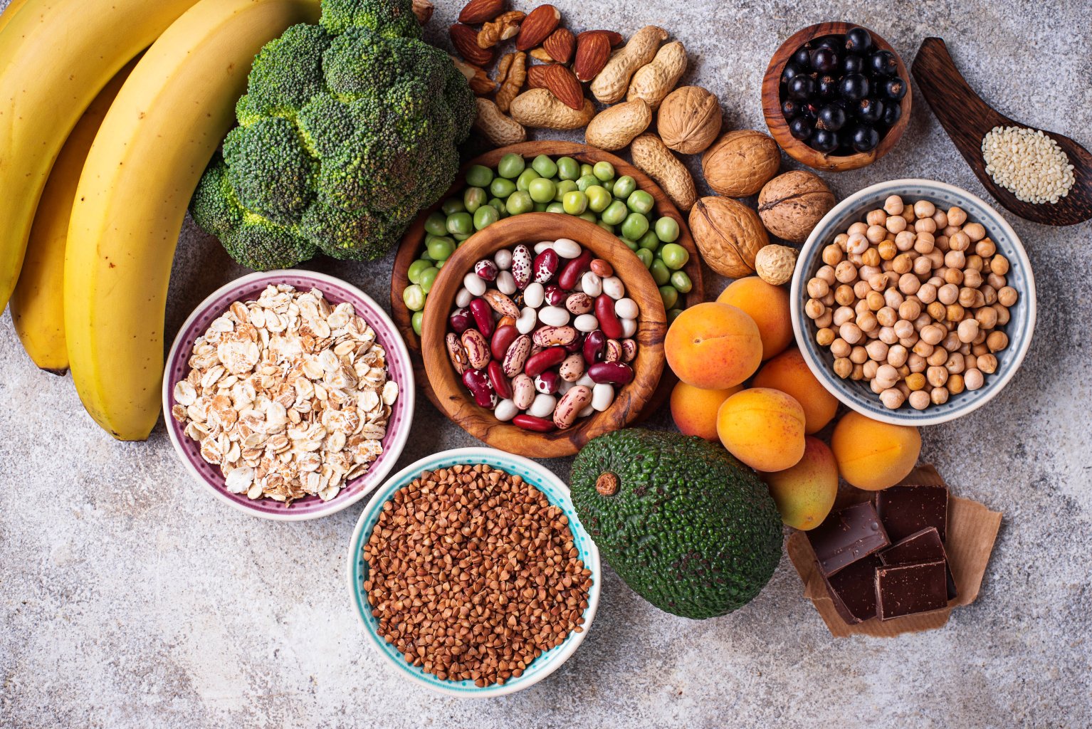 Eine Vielzahl gesunder Lebensmittel wie Bananen, Brokkoli, N&uuml;sse, Samen, Erbsen, Bohnen, Hafer, Kichererbsen, getrocknete Aprikosen, Avocado, Buchweizen und dunkle Schokoladenst&uuml;cke auf einer hellen Unterlage.