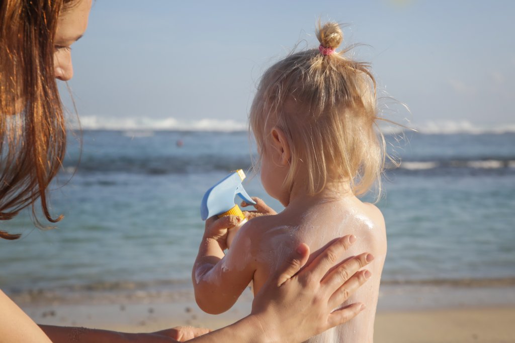 Eine Frau tr&auml;gt am Strand Sonnenschutz bei Kindern auf den R&uuml;cken eines kleinen Kindes auf. Das Kind, das eine Sonnenschutzflasche in der Hand h&auml;lt, blickt auf das Meer, w&auml;hrend die Wellen anrollen und sich der blaue Himmel &uuml;ber ihm ausbreitet.